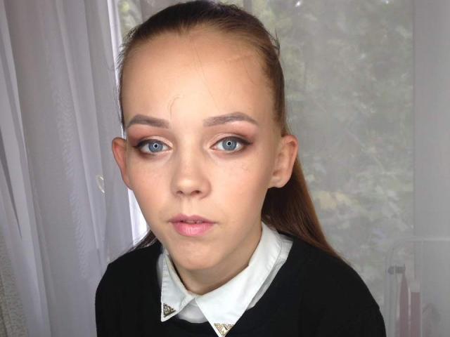 JeannieTina has pale skin, blue eyes, and brown hair in a ponytail. She's wearing a black top with a white collar and gold buttons, looking serious.