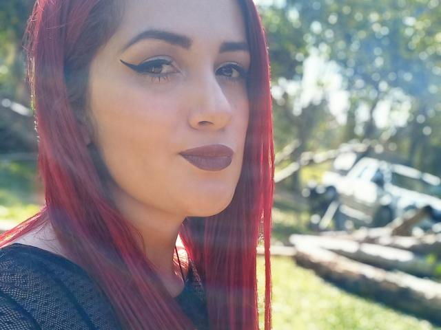 BrunaRousse69, con cabello rojo, cejas pobladas, delineador de ojos alado y labial morado oscuro, vestida con una blusa negra, mirando a la cámara con una ligera sonrisa. Luz solar en el fondo..