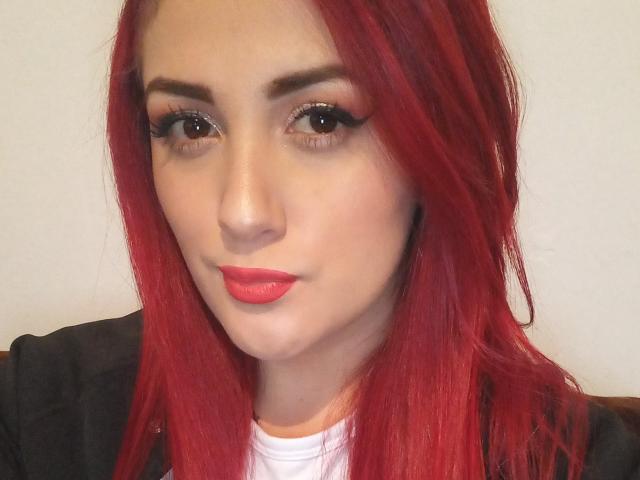 BrunaRousse69 a du rouge à lèvres rouge, un crayon à paupières rouge, un fard à paupières rouge et des cheveux roux. Elle regarde l'appareil photo avec ses yeux marron, et porte une veste noire par-dessus une chemise blanche..