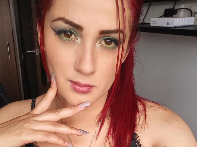 BrunaRousse69 con cabello rojo, sombra de ojos verde y dorada, ojos marrones, labios rosados y uñas largas rosadas, tocándose la barbilla, vistiendo una blusa negra de encaje, mirando a la cámara..