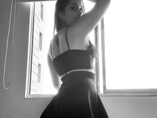 BrunaRousse69, con la schiena rivolta verso la fotocamera, che alza un braccio, indossa un top e una gonna senza spalline, con i glutei leggermente sollevati, in una foto in bianco e nero..