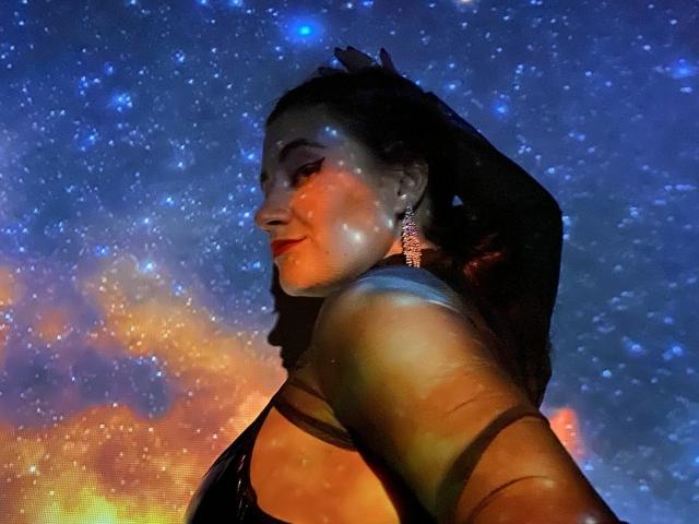 ArleenHunter with dark hair, glowing skin, and red lipstick, wearing a black top, showing her back and side, with starry lights on her and wearing elegant earrings.