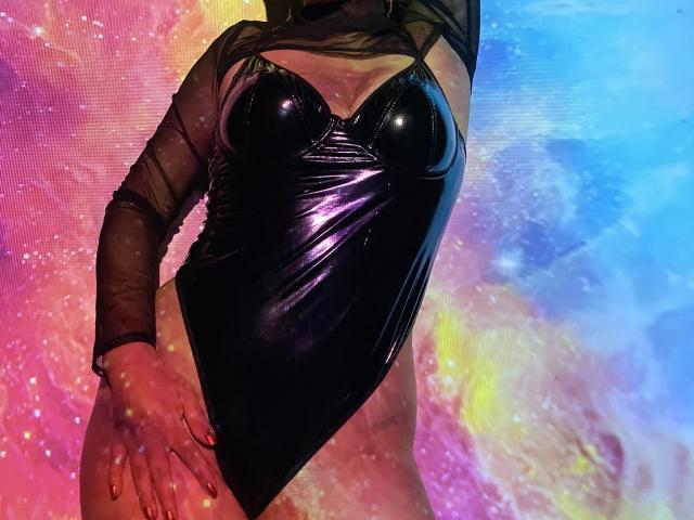 ArleenHunter wearing a shiny black leather bodysuit with cutout chest and mesh sleeves, standing with hands on hips, highlighting her tits and slim waist.