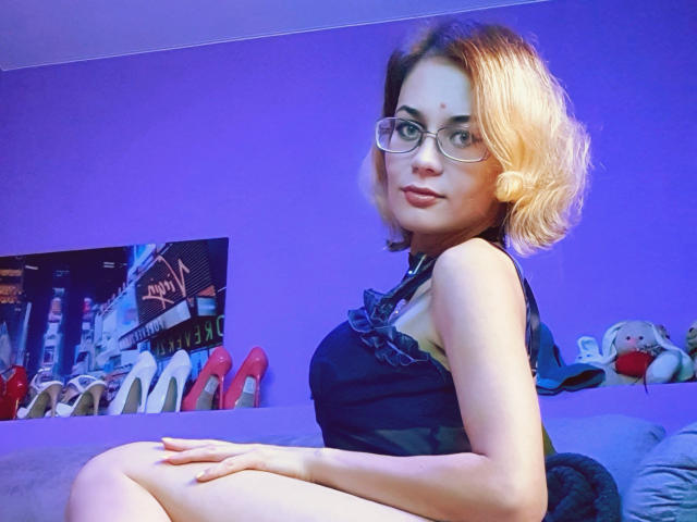 MilanaPure with blonde, wavy hair, glasses, and a black dress, sitting on a couch, showing her ass, looking over her shoulder with a cute smile.