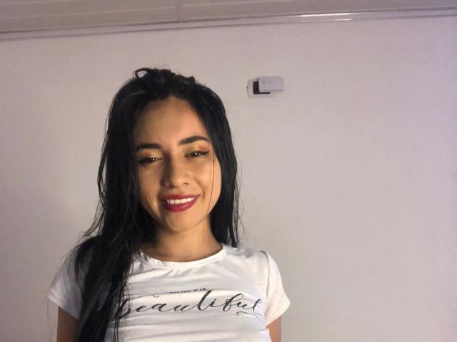 ElizabethRosh, com cabelos pretos longos, olhos castanhos e batom rosa, vestindo uma camisa branca, sorrindo, mostrando os seios e com um rosto bonito e sexy..