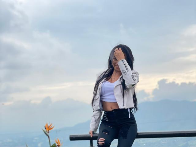 Elizabeth Rosh mit langem, schwarzem Haar, die ein weißes Crop-Top, eine graue Jacke und zerrissene schwarze Jeans trägt, sitzt auf einer Bank, wobei sie sich die Hand vor den Mund hält. Im Hintergrund ist ein bewölkter Himmel zu sehen..