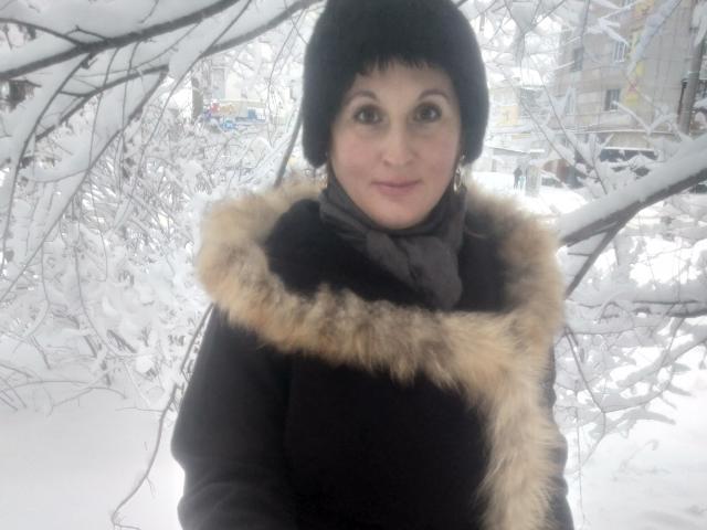 KelliRedhaer, con un corte de pelo bob negro, vistiendo un abrigo negro con detalles de piel, una bufanda gris, de pie en una zona nevada, con ramas sobre su cabeza, sonriendo ligeramente, ojos marrones, piel clara, pequeños aros..