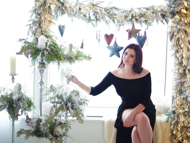 SweetAdriana, with red hair and off-shoulder black dress, sits on white chair, touching silver candleholder, surrounded by greenery, white candles, and golden star and heart ornaments.