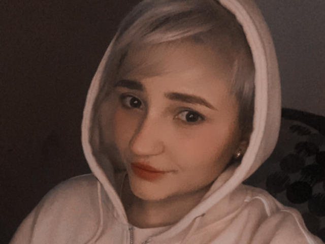 WendyMoira69 mit kurzen, weißen Haaren, die einen weißen Kapuzenpullover trägt, hat ein süßes Gesicht, rote Lippen und eine kleine Nase und blickt mit einem leichten Lächeln in die Kamera..