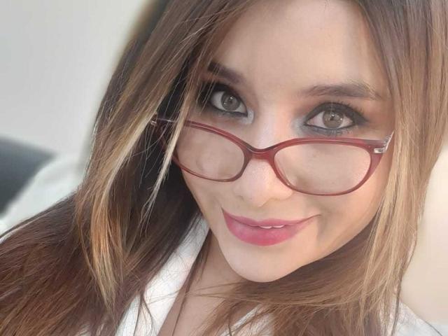 Sharlote Durant, con i capelli castani, gli occhiali rossi e il rossetto rosa, sorride alla fotocamera con gli occhi grigi. Ha un viso carino e attraente, e una leggera abbronzatura..