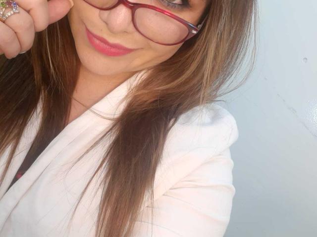 Sharlote Durant, avec de longs cheveux bruns, des lunettes rouges, du rouge à lèvres rose et une chemise blanche, tenant ses lunettes avec un sourire discret, montrant sa bague..
