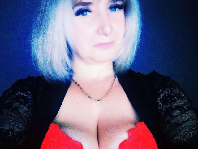 VAlica with short blonde hair, blue eyes, and a slight smile, wearing a black lace top and red lace bra, showing large tits. She's also wearing a gold chain.