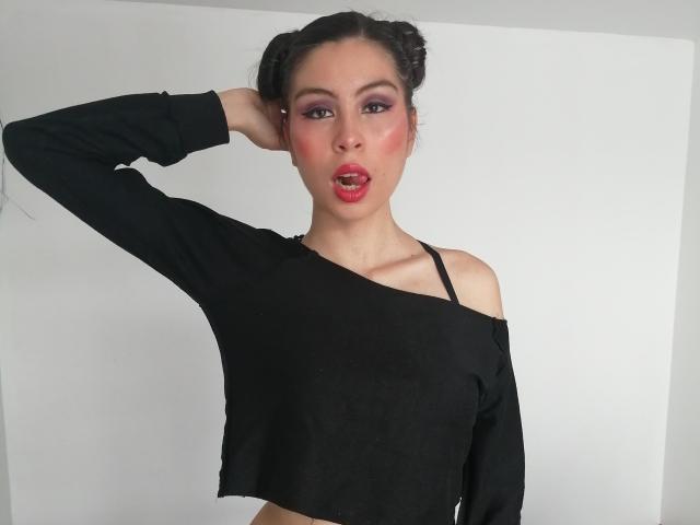 IsaTimeSweet, avec ses cheveux foncés tressés, un haut noir à une épaule, un fard à paupières rose, un rouge à lèvres rouge, et un bandeau de soutien-gorge noir visible, une main dans les cheveux, regardant la caméra, la bouche légèrement ouverte..
