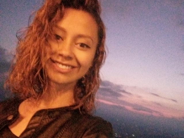 ElizaJones with wavy brown hair, smiling, showing teeth, wearing a black shirt, has a cute face and small tits, in a slightly blurry selfie at sunset.