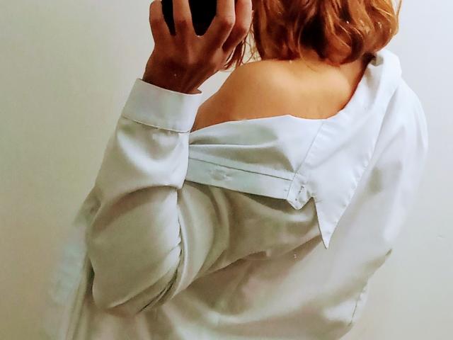 ElizaJones with wavy red hair, off-shoulder white shirt, showing bare shoulder, holding black phone, shirt button partially undone, right arm raised, white wall background.