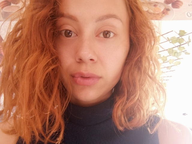 Eliza Jones, com cabelo ondulado, avermelhado-laranja, pele clara, olhos castanhos e lábios rosados, vestindo um suéter preto de gola alta, olhando para a câmera com uma expressão neutra..