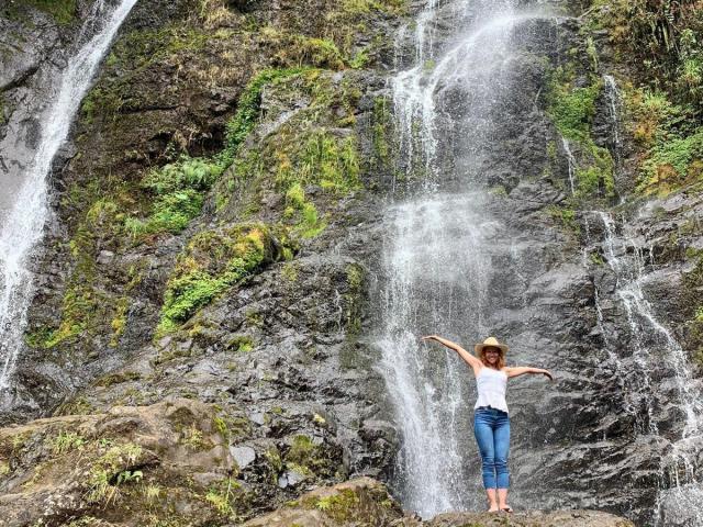 Eliza Jones está de pie bajo una cascada, con los brazos extendidos, vistiendo una camiseta blanca y pantalones vaqueros azules, con un sombrero de sol, y agua salpicando a su alrededor, en un terreno rocoso y cubierto de musgo..