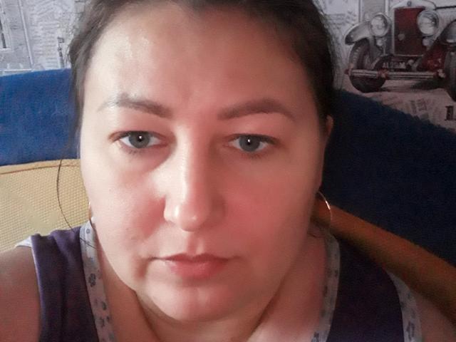 Riniya, con labios rosados y voluminosos, una nariz pequeña y ojos marrones, vestida con una blusa morada y gris, tiene un rostro ligeramente rechoncho y cabello castaño oscuro recogido..