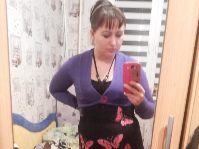 Riniya, con i capelli castani corti, un cardigan viola, una maglia nera in pizzo e una gonna rosa a forma di farfalla, sta scattando un selfie allo specchio, mostrando il seno, in piedi con le mani sui fianchi..