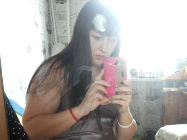 Riniya, com cabelos longos e escuros, vestindo uma blusa cinza sem mangas, tira uma selfie no espelho com um celular rosa, com a luz refletindo em sua testa, e um bracelete vermelho no pulso direito..