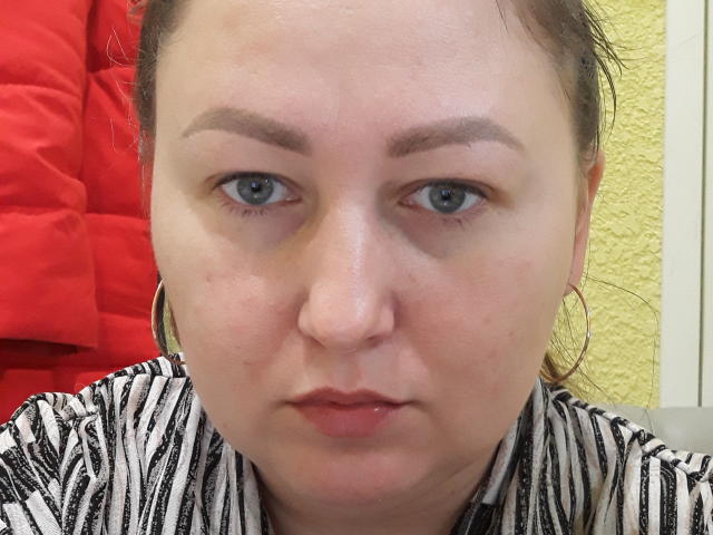 Riniya tem um rosto gordinho, olhos azuis e lábios vermelhos levemente inchados. Ela está vestindo uma camisa listrada e grandes brincos de argola, e ao fundo, há um casaco vermelho e volumoso..