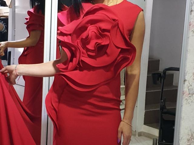 TheSunshine porte une robe rouge avec un grand tissu plissé, ressemblant à des roses, sur la poitrine, et tient le côté avec une main. Son reflet est visible dans un miroir..