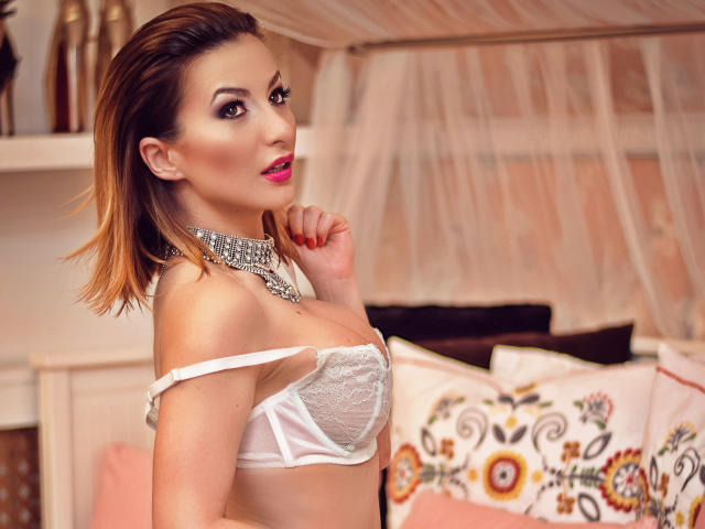 MayaEva with medium tits, wearing a white lace bra, silver necklace, and pink lipstick, looking over her shoulder with a hand near her mouth, in a bedroom setting.
