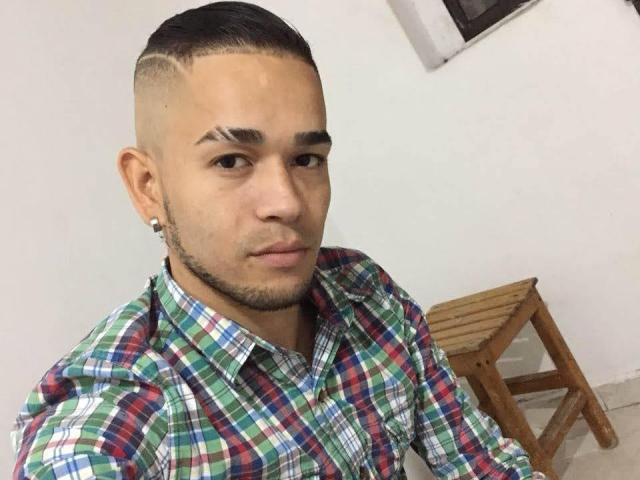 JhonnyThePaisa ha un taglio di capelli rasati su un lato, con sfumature, orecchini piccoli e indossa una camicia a quadri verde, rossa e blu, con un'espressione seria..