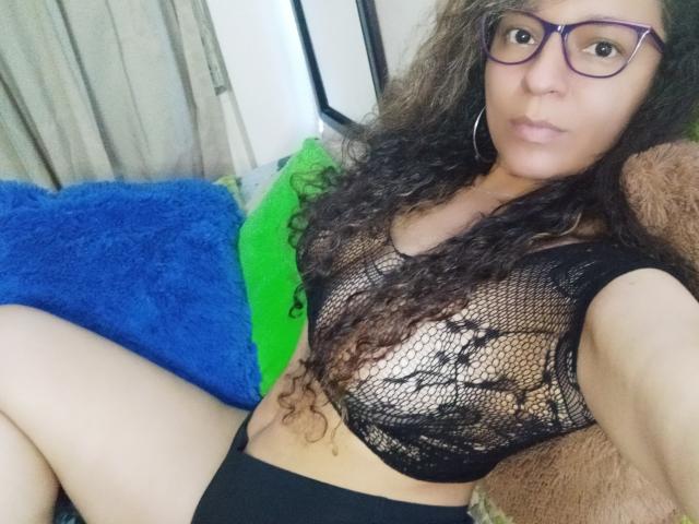 Angelonix with curly brown hair, purple glasses, and hoop earrings, wearing a black lace top, showing her tits, black panties, and a serious expression, sitting on a blue pillow.