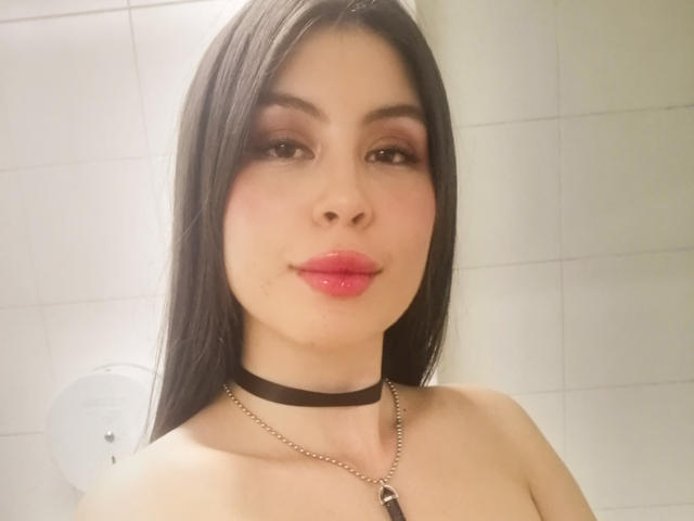 IsaTimeSweet tiene el pelo largo y negro, labios rosados y brillantes, maquillaje de ojos marrón y ahumado, un collar negro y un collar de plata con un colgante negro, mostrando los hombros descubiertos..
