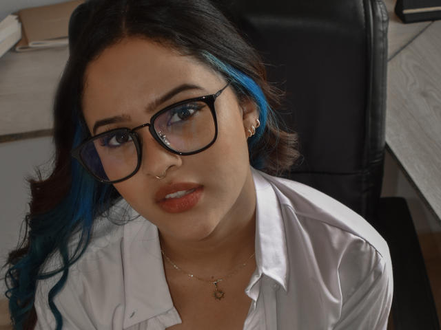 Stephanie Valdez mit blau gespitzt, Brille, Nasenring und weißem Hemd, die nach oben schaut, trägt eine Halskette mit einem kleinen Herzanhänger und hat den Mund leicht geöffnet..