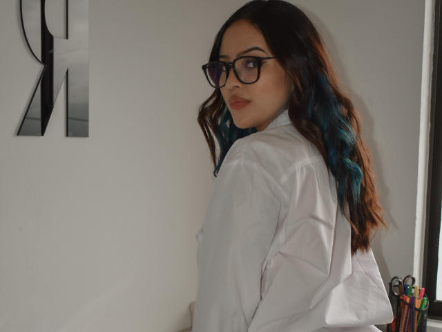 StephanieValdez with blue ombre hair, black glasses, and white shirt, looking over her shoulder, showing her ass. She has a slight smile and a cute, sexy look.
