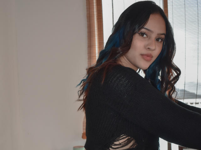 Stephanie Valdez, con capelli ondulati castano-blu, indossa una maglia a maglia nera, guarda oltre la spalla con un leggero sorriso, mostrando il piercing al naso e un trucco leggero..