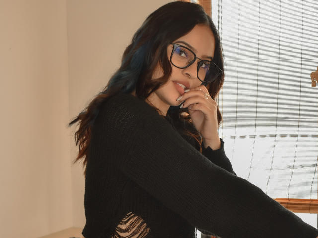 Stephanie Valdez, con cabello oscuro y ondulado, gafas negras y una blusa de punto negra, mordiéndose el dedo, mostrando un anillo y con una expresión linda, ligeramente seductora..