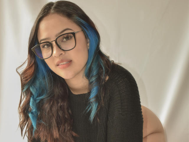 Stephanie Valdez, con riflessi blu, occhiali neri e maglione nero, è china in avanti, mostrando il sedere scoperto, e sorride alla telecamera. Luce soffusa, capelli leggermente ondulati, carina, sexy..