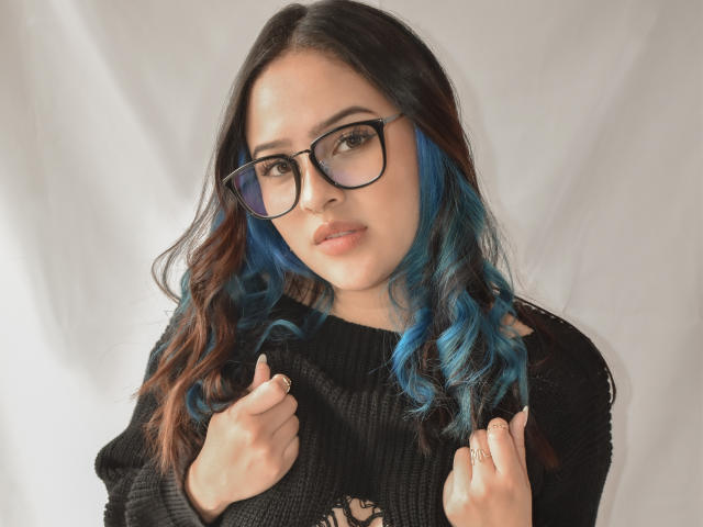 Stephanie Valdez, con capelli ondulati castano-blu, indossa occhiali neri e un maglione nero, tiene il maglione tra le mani, mostrando un anello al dito destro..