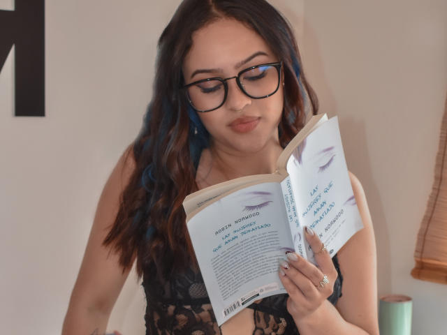 Stephanie Valdez, con i capelli scuri e ondulati, riflessi blu, occhiali neri e un reggiseno di pizzo nero, sta leggendo un libro, concentrata sulle pagine, appare carina, ed è in piedi in una stanza con una luce soffusa..
