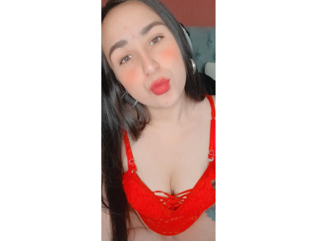 OriianaT, con cabello negro largo, vistiendo un sujetador de encaje rojo, con labios rojos entrecerrados y grandes aretes de plata, mostrando sus pechos. Está mirando hacia arriba con una expresión linda y juguetona..