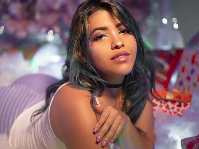KimLafayette, avec ses cheveux bleu-gris, sa robe blanche et son collier, allongée sur un oreiller à rayures violettes, regardant séductrice l'appareil photo, avec du rouge à lèvres rose, des yeux violets et un vernis à ongles rose, éclairage doux..
