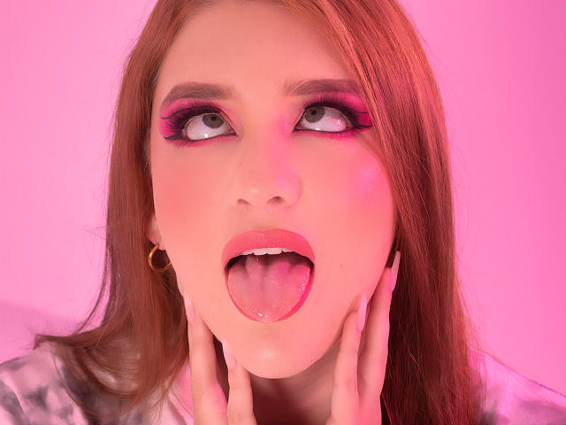 Das Gesicht von MelisaLee mit rosigem Make-up, die Zunge herausgestreckt, die Hände auf den Wangen, blickt nach oben. Sie hat rotes Haar, rosafarbenen Lippenstift und große Ohrringe. Ihre Augen sind weit geöffnet und sie wirkt attraktiv..