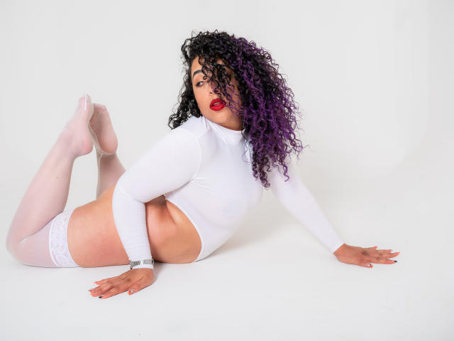 Scarlett, avec ses cheveux bouclés violets, ses lèvres rouges, son crop top blanc à manches longues, ses bas blancs montants jusqu'aux cuisses et son bracelet, allongée sur un fond blanc, les bras et les jambes écartés, montrant ses fesses..