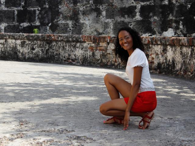 MeganGil est accroupie, les fesses en arrière, elle porte un haut blanc et un short rouge, et elle sourit à la caméra. Elle a les cheveux noirs et bouclés et porte des sandales rouges..