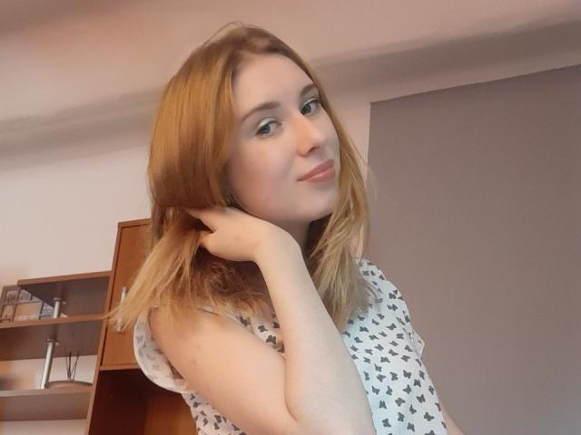 MaudieMel, com cabelo castanho claro, vestindo uma camisa branca estampada, segurando o pescoço, sorrindo suavemente, pele clara, olhos azuis, corpo de estatura média, maquiagem leve, em ambiente interno, selfie casual..