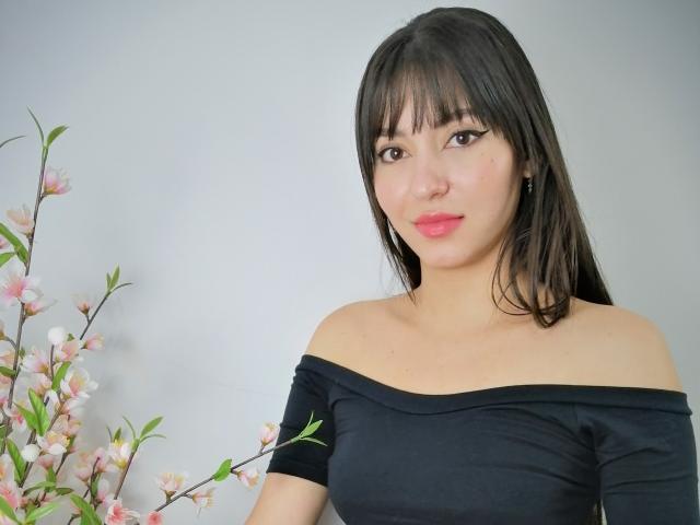 NalaAndDrako, donna asiatica con lunghi capelli neri, indossa una maglietta nera con una spalla scoperta, rossetto rosa, piccoli orecchini, tiene dei fiori di ciliegio, un sorriso dolce, pelle chiara, leggero trucco, viso carino..