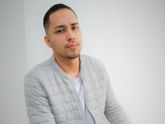 NalaAndDrako, short-haired dude with a light beard, wearing a grey quilted jacket over a white tee, looking slightly confused or bored.