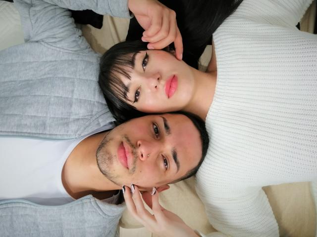 NalaAndDrako, con i capelli scuri, le labbra rosa e un maglione bianco, sdraiata accanto a Drako, con i capelli neri corti, una leggera barba e una felpa grigia, entrambi che guardano la fotocamera. La mano di NalaAndDrako sul viso di Drako..