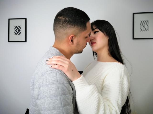 NalaAndDrako con cabello negro corto, camisa gris, y NalaAndDrako con cabello negro largo, suéter blanco, tocándose las caras, mirándose a los ojos, expresiones suaves, momento íntimo..