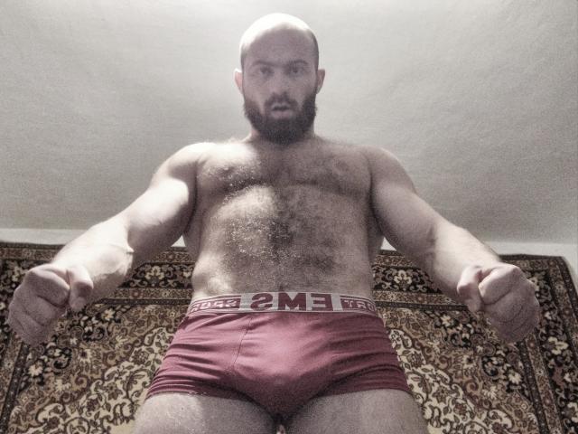 AlanStrongs, chauve, barbu, musclé, avec une poitrine poilue, portant des sous-vêtements rouges, poings en avant, regardant vers le bas, expression sérieuse, debout sur un tapis orné, prise de vue en contre-plongée..