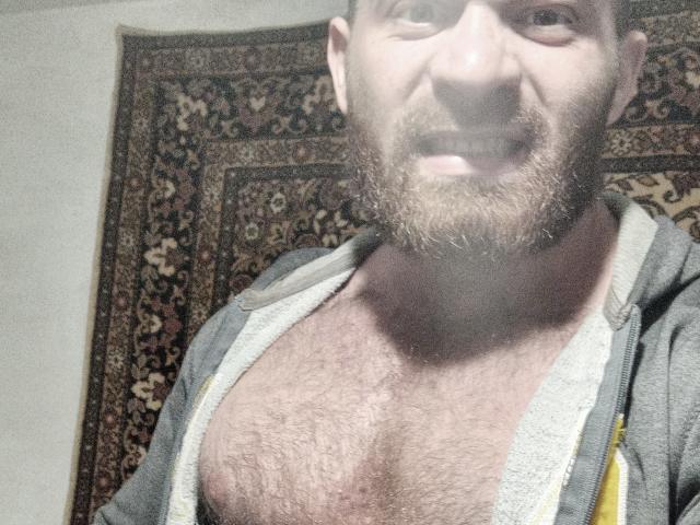 AlanStrongs, ein Mann mit Bart und behaarter Brust, trägt eine graue, aufreißbare Kapuzenjacke, die offen ist, um seine behaarte, muskulöse Brust zu zeigen. Er lächelt leicht, und das Bild ist ein Selfie von seinem Kopf und Oberkörper..