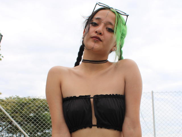 SalomePrince, avec des cheveux tressés verts et noirs, un haut noir sans bretelles avec découpes, un collier, un anneau au nez et un anneau aux lèvres, portant des lunettes de soleil sur la tête, regardant vers le bas..